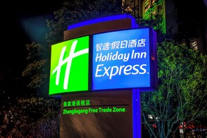 Une enseigne Holiday Inn Express aux couleurs vert et bleu, située dans une zone commerciale, avec des arbres et des bâtiments en arrière-plan.