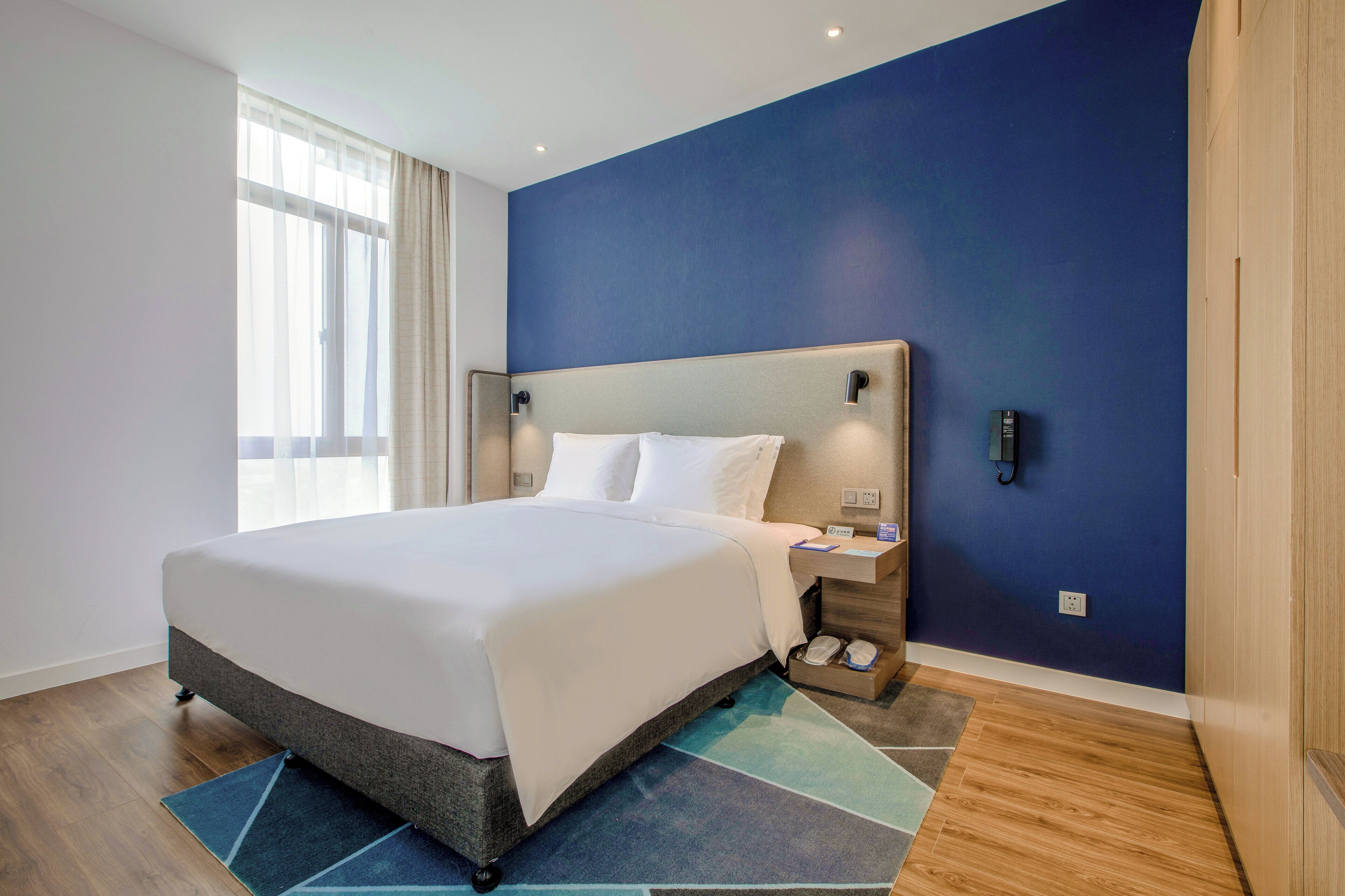 Habitación de hotel con una cama grande, una mesita de noche, teléfono en la pared y una ventana con cortinas translúcidas.
