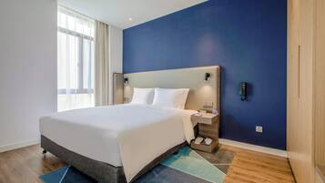 Habitación de hotel con una cama grande, una mesita de noche, teléfono en la pared y una ventana con cortinas translúcidas.