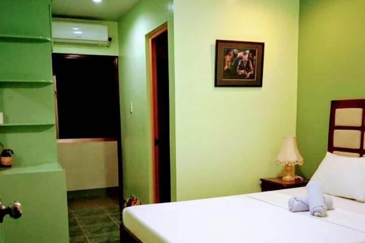 A room with a bed, a nightstand, a lamp, a framed picture on the wall, and an air conditioning unit.