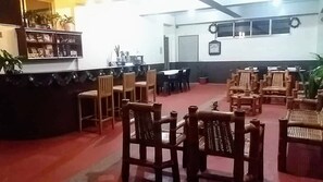 Un hall d’entrée avec un comptoir de réception, plusieurs chaises en bois, un coin repas avec des tables et des chaises, et un tapis rouge.