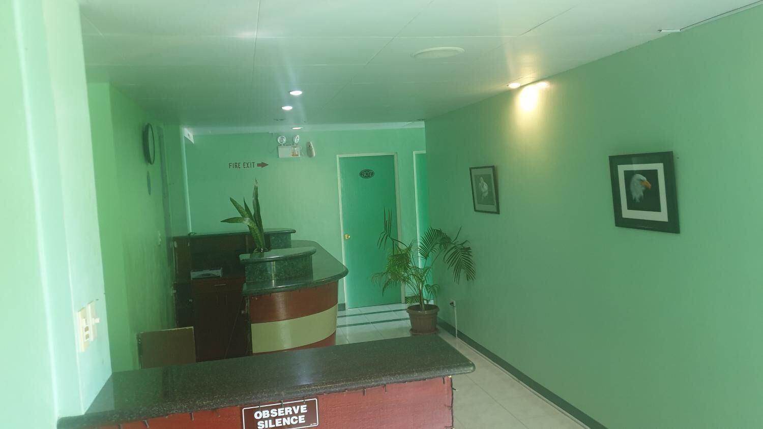 A reception area with a counter, a sign instructing to observe silence, a door marked 'EXIT', and potted plants.