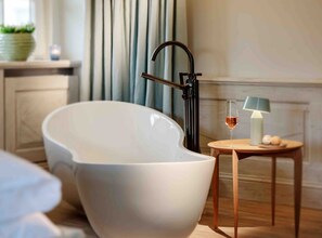 A modern bathroom with a freestanding bathtub, a small wooden side table with a lamp, and a glass of wine.