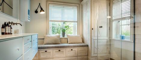 A bathroom with a wooden floor, a bench with pillows, a glass shower enclosure, and a window with blinds.
