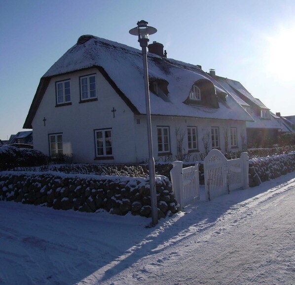 A snow-covered house with a stone wall, a wooden gate, and a street lamp.