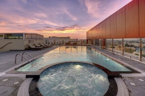 A rooftop swimming pool with a view of the city at sunset, surrounded by lounge chairs and a modern building.