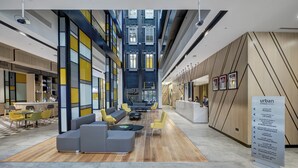Een moderne hotellobby met een hoge plafond, een centraal zithoek met banken en tafels, een receptiebalie, en een bord waarop de faciliteiten staan.