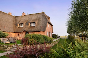 A thatched-roof house with a brick exterior, a stone pathway, and a garden with shrubs and a lawn.