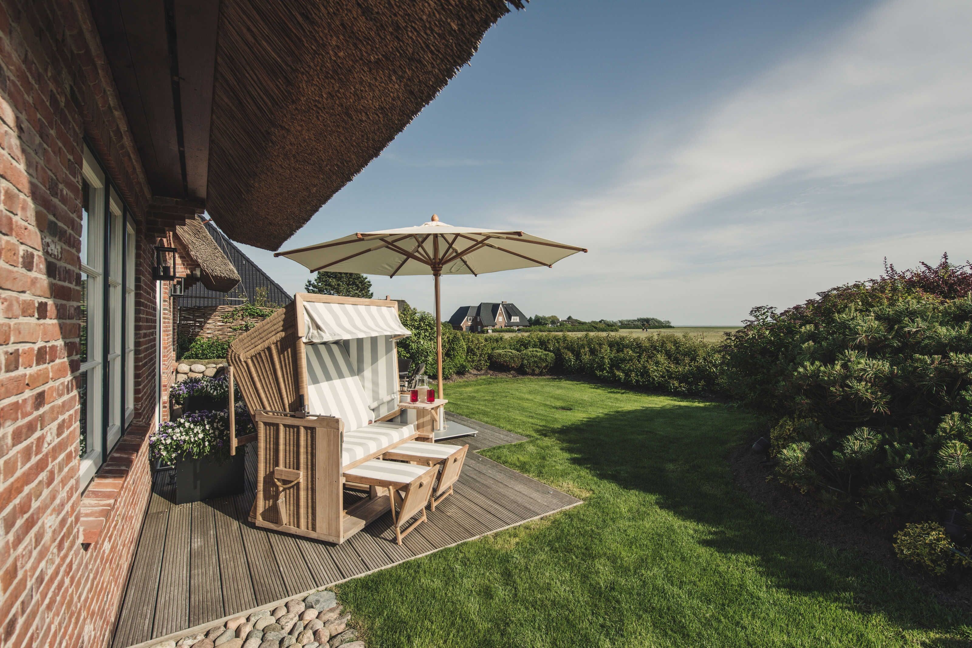 Una terraza de madera con techo de paja, un área de estar al aire libre con cojines y vistas a un jardín cuidado.