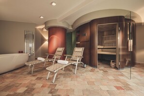 Ein Wellnessbereich mit Badewanne, drei Liegestühlen und einer Saune mit Glasfront.
