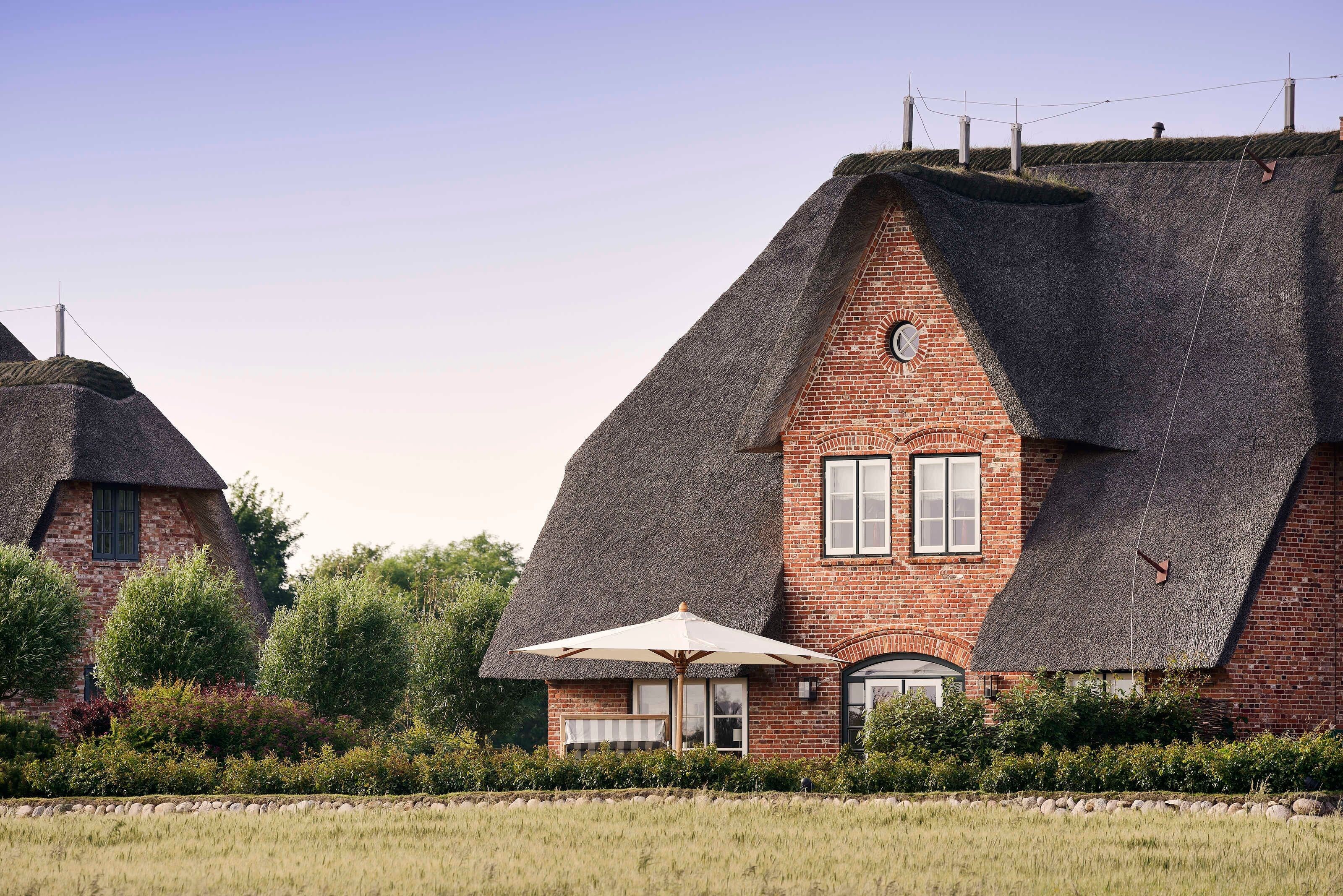 A traditional thatched-roof house with a brick lower section, surrounded by a well-maintained garden and a field.
