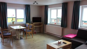 Ein Wohnzimmer mit Sofa, Couchtisch, Fernseher auf einem Schrank, Esstisch mit Stühlen und einem Fenster mit Vorhängen.