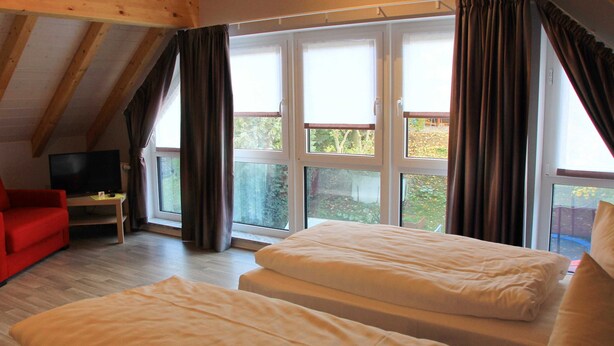 Una habitación con dos camas, un sofá rojo, un televisor y ventanas grandes con cortinas.