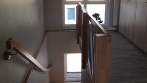 Una escalera de madera con barandilla que lleva a un rellano. Se ve una ventana con persianas en el fondo.