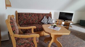 En stue med en blomstret sofa, træarmstole, et lille træbord med en vase, et fjernsyn på et stativ og en grøn skammel.