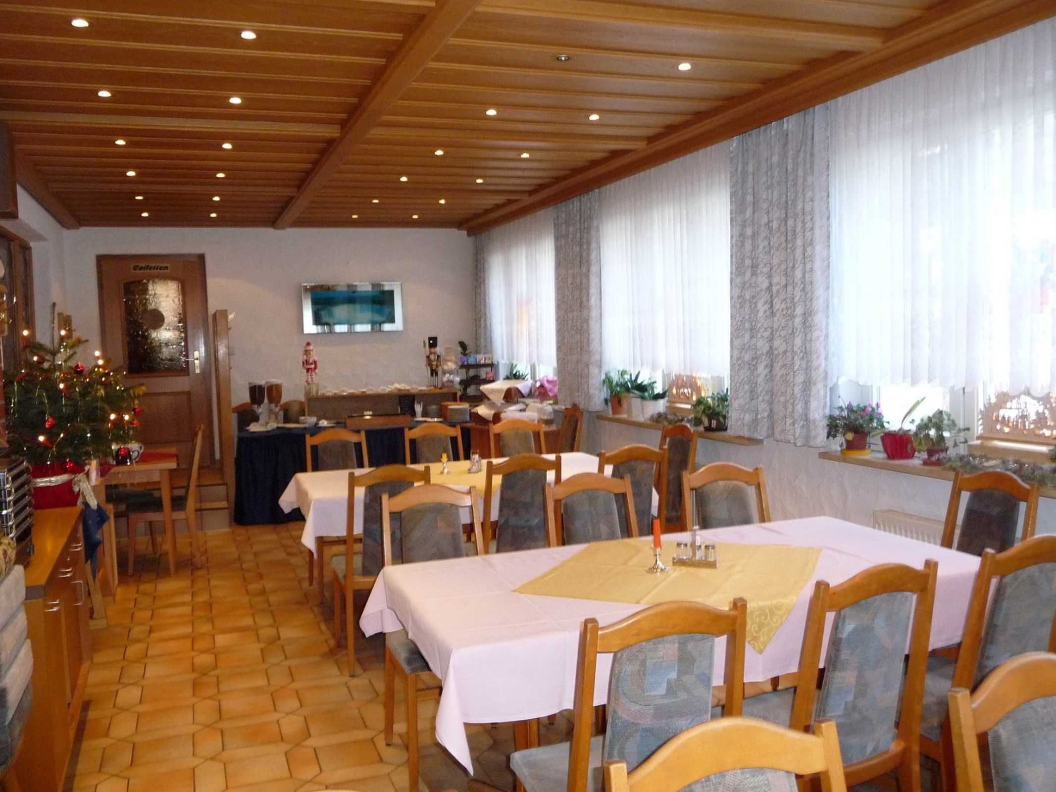 Un espace repas avec des tables et des chaises en bois, un sapin de Noël et une grande fenêtre avec des rideaux.