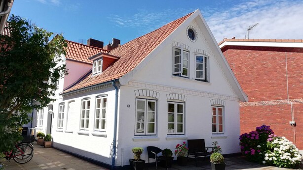 A white building with a red-tiled roof, multiple windows, and a small porch area with potted plants and a bench. - Carl-Häberlin-Str. 15, EG (Wyk auf Föhr)