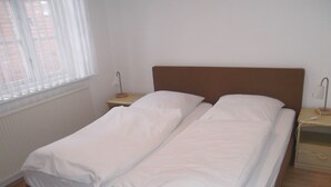 A twin bed with white bedding in a room with a wooden floor, a window with blinds, a bedside table with a lamp, and a radiator. - Carl-Häberlin-Str. 15, EG (Wyk auf Föhr)