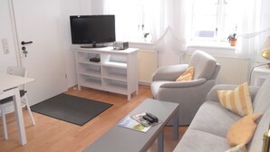 A living room with a TV on a white stand, a grey sofa, a small table, and a white chair. - Carl-Häberlin-Str. 15, EG (Wyk auf Föhr)
