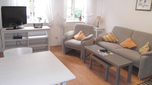 A living room with a TV, a sofa, a chair, a coffee table, and a window with curtains. - Carl-Häberlin-Str. 15, EG (Wyk auf Föhr)