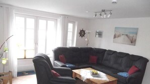 Een woonkamer met een zwarte bank, een houten salontafel en een stoel. Er is een raam met gordijnen, een wandklok en een schilderij.