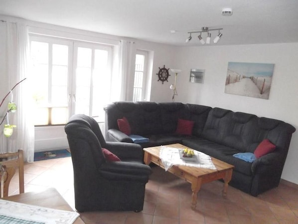A living room with a black sofa, a wooden coffee table, and a chair. There is a window with curtains, a wall clock, and a painting.