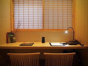 Un espace de travail comprenant un bureau, deux chaises, une fenêtre avec des stores, une lampe et quelques livres.