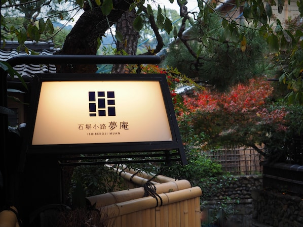 Une enseigne de style japonais traditionnel, ornée d’un logo et d’un texte, se détachant sur un fond de verdure luxuriante et de feuillage automnal.