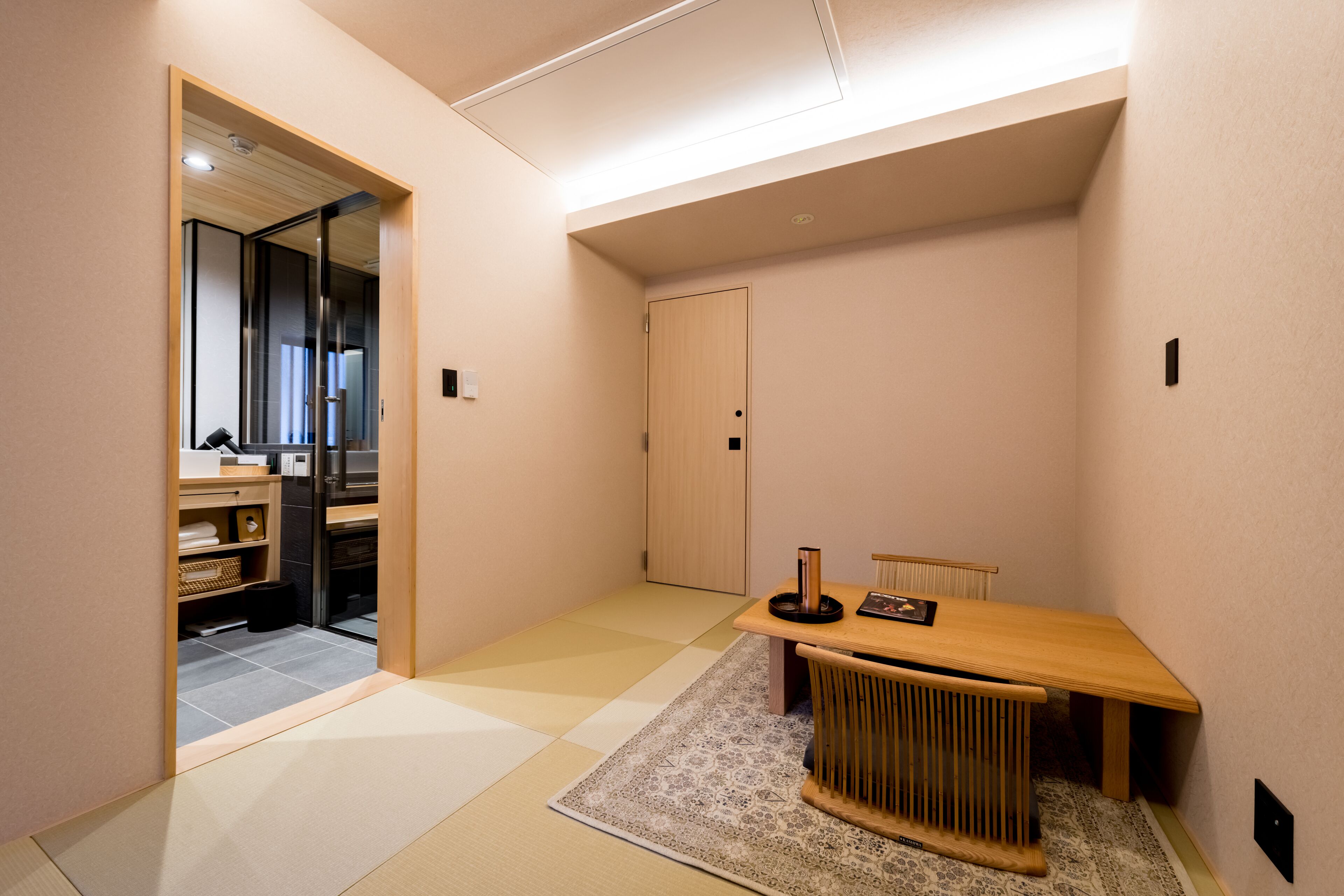 A modern, minimalist room with a wooden table and chairs, a small rug, and a sliding door leading to a kitchen area.