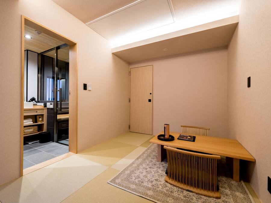 A modern, minimalist room with a wooden table and chairs, a small rug, and a sliding door leading to a kitchen area.
