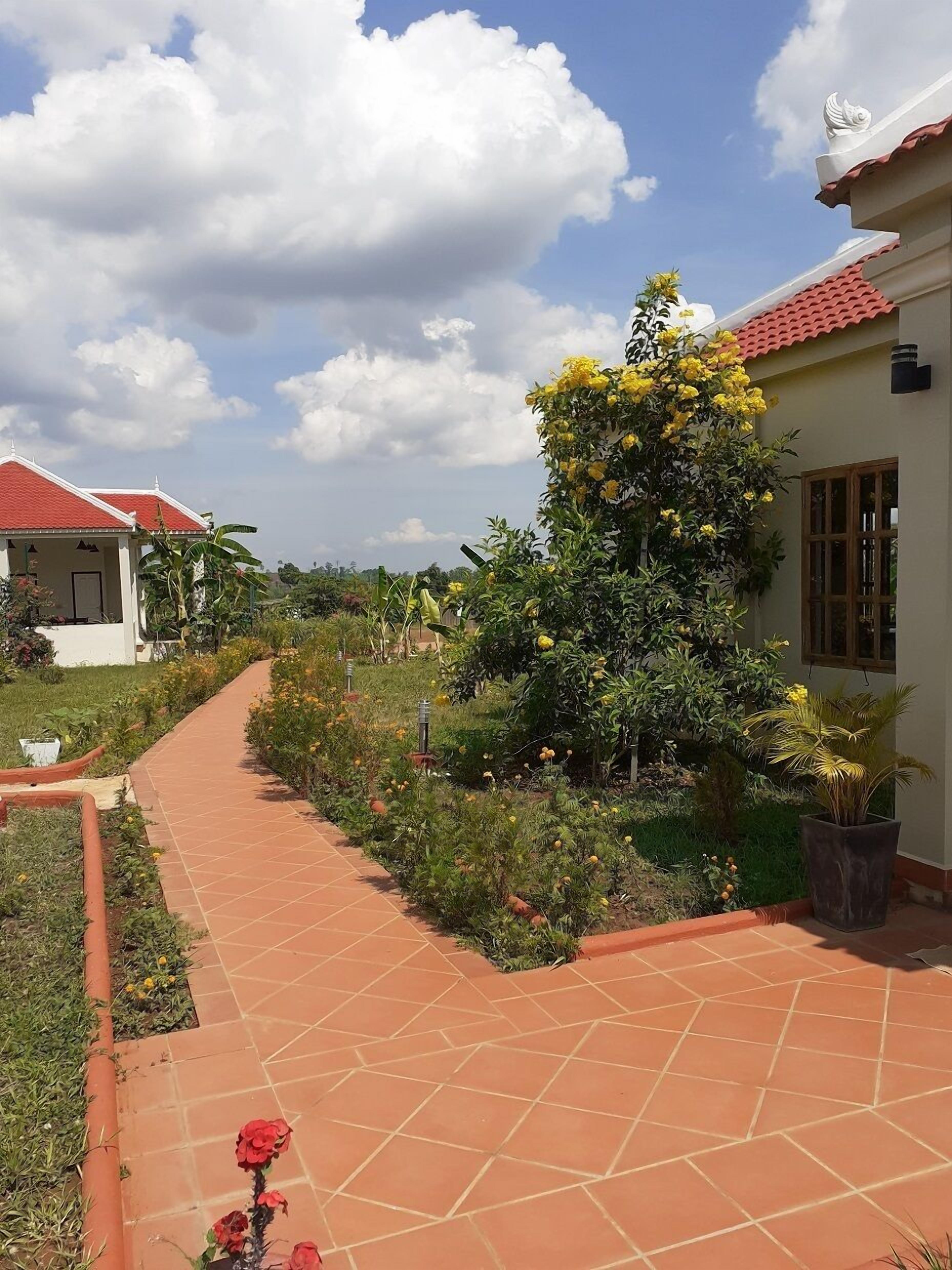 A pathway leading to a building with a red-tiled roof, surrounded by lush greenery and flowering plants.