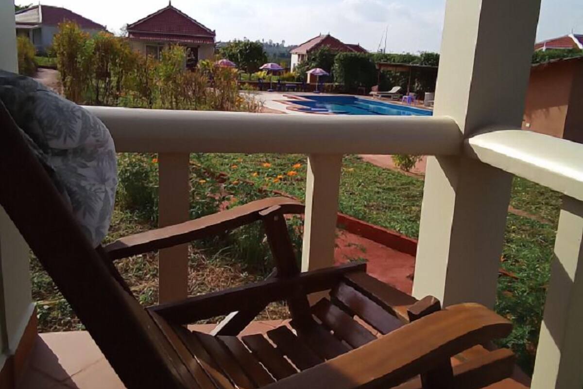 A wooden chair on a balcony overlooking a swimming pool with a garden and houses in the background.