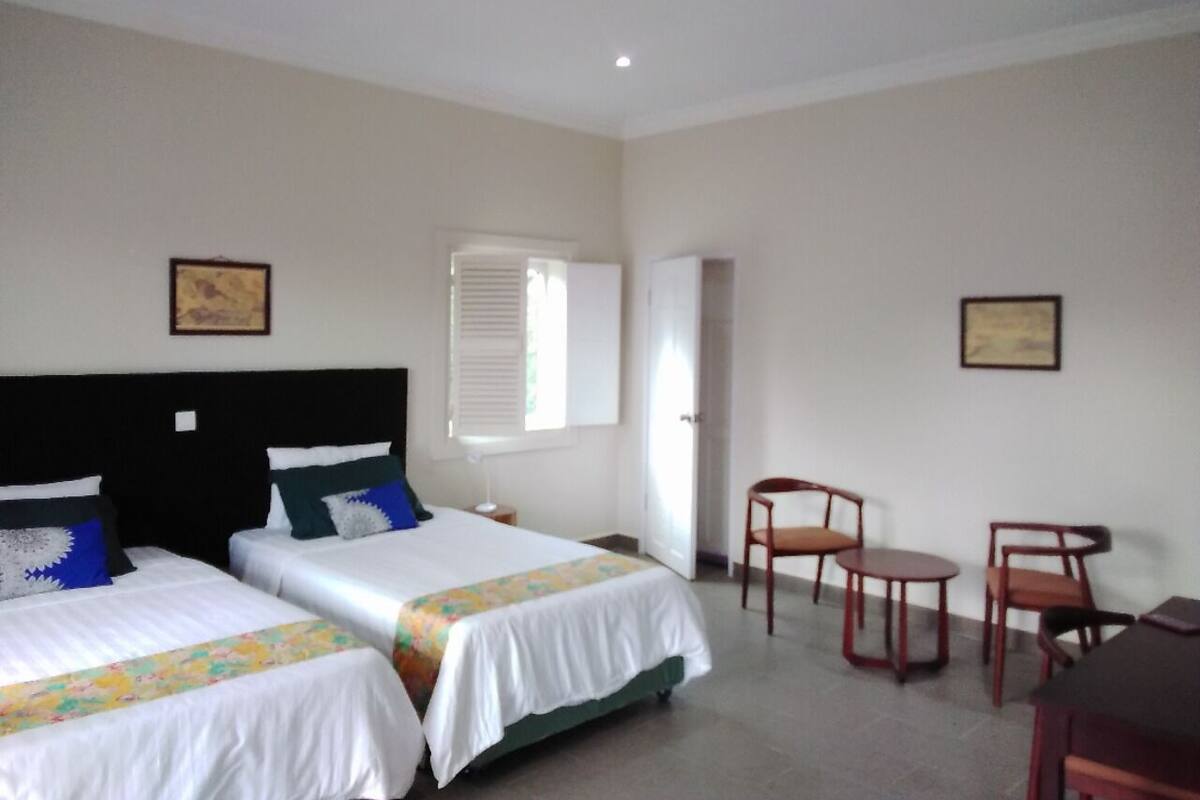 A hotel room with two beds, a ceiling fan, a window with blinds, and a small table with chairs.