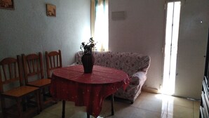 A dining area with a round table covered in a red tablecloth, surrounded by wooden chairs. A vase with flowers is placed on the table. There is a patterned sofa against the wall and a window with curtains. - Cozy and central bass (Ciudad Real)