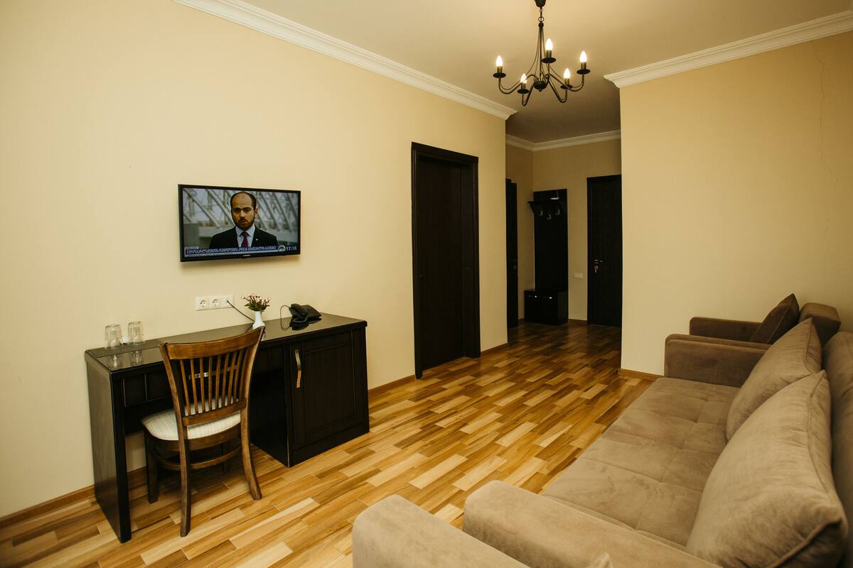 A living room with a wooden floor, a sofa, a dining table with chairs, a TV mounted on the wall, and a chandelier.