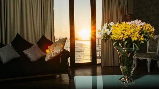 A room with a sofa, a vase of flowers, and a view of the sunset over the water. - Kobuleti Pearl Of Sea Hotel & Spa (Kobuleti)