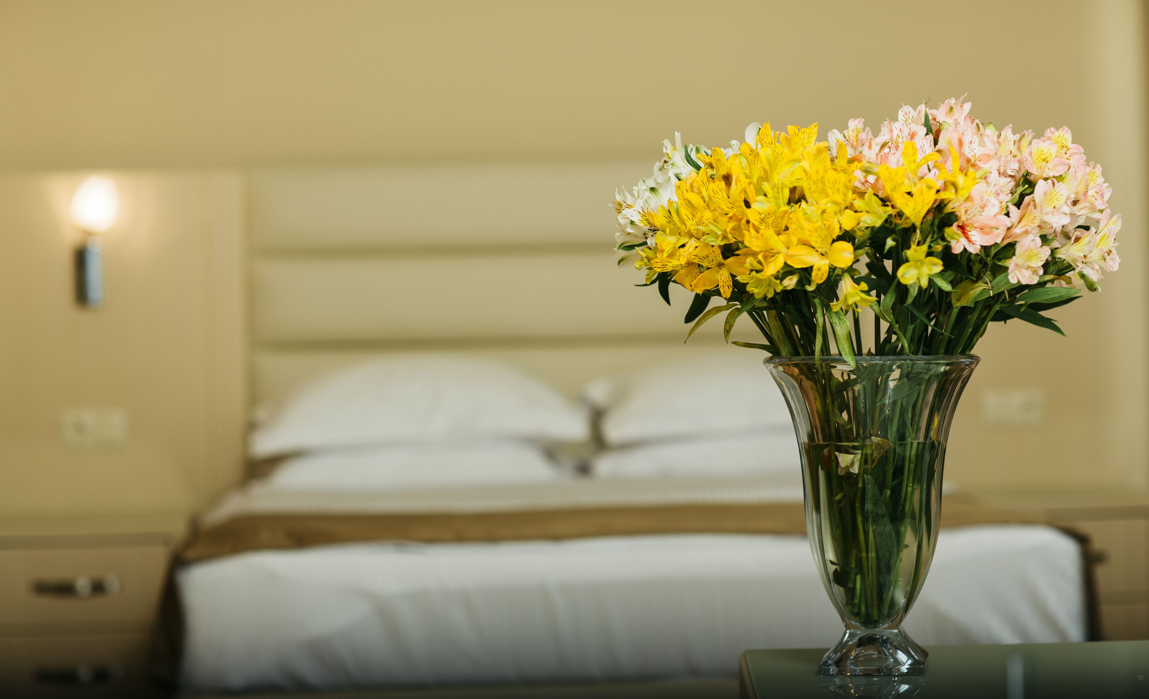 A bed with white linens and a vase with yellow and pink flowers on a bedside table.
