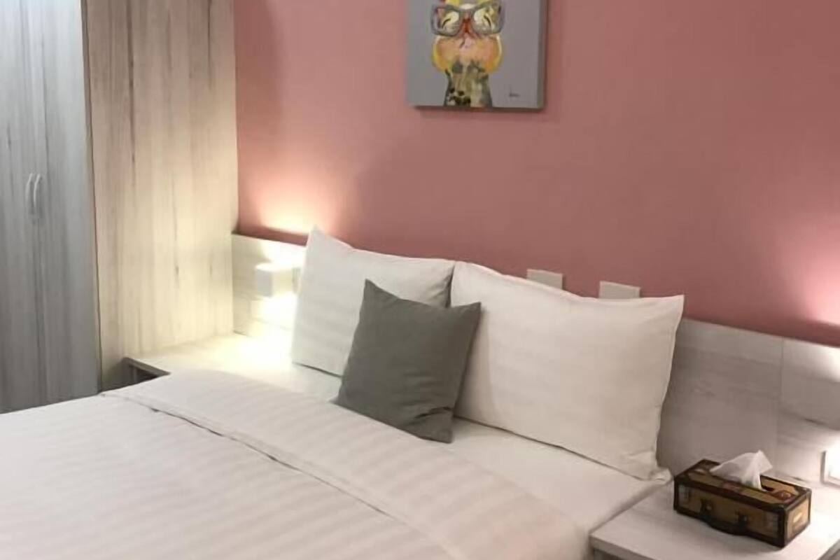 A hotel room with a bed, pillows, a nightstand, a painting, and a wardrobe.