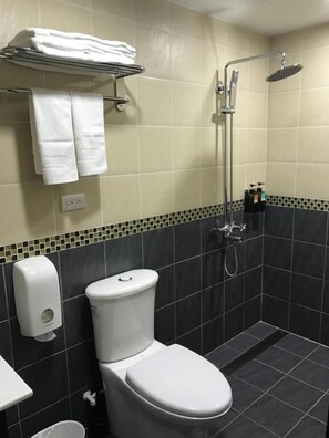 A bathroom with a toilet, a hand dryer, a towel rack with towels, a showerhead, and a tiled floor.