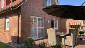 A brick house with a large glass door, a covered patio with wicker furniture, and a barbecue grill. - North Sea vacation with children (Dornum)