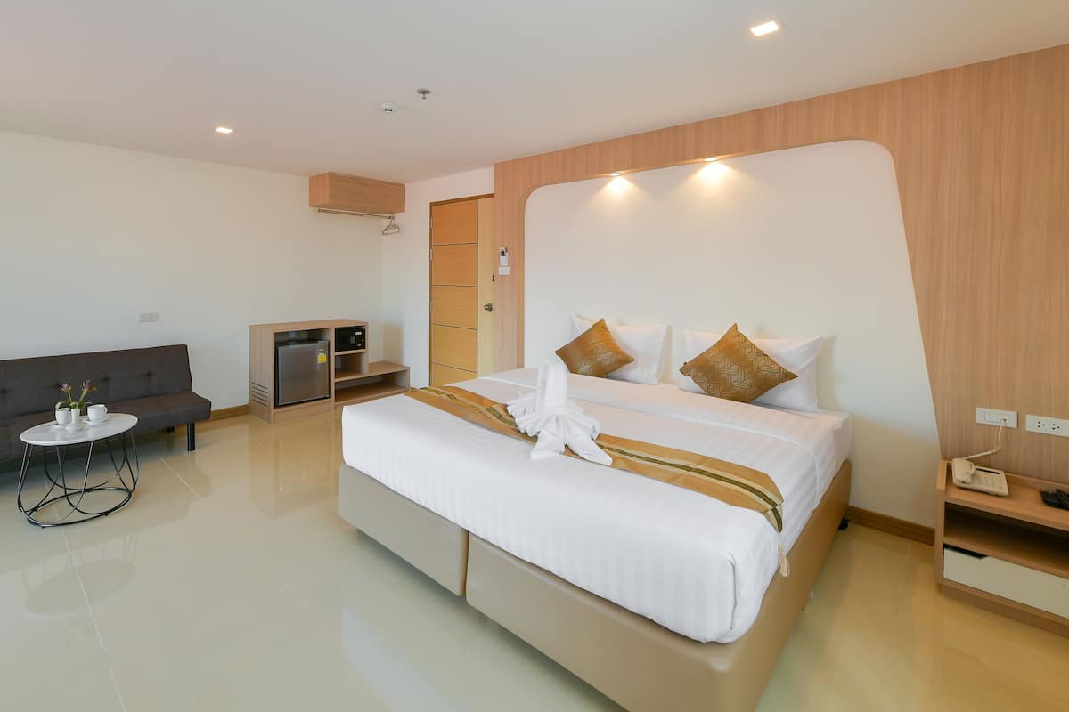 A modern hotel room with a large bed, a sofa, a small table, a minibar, and a wooden accent wall.
