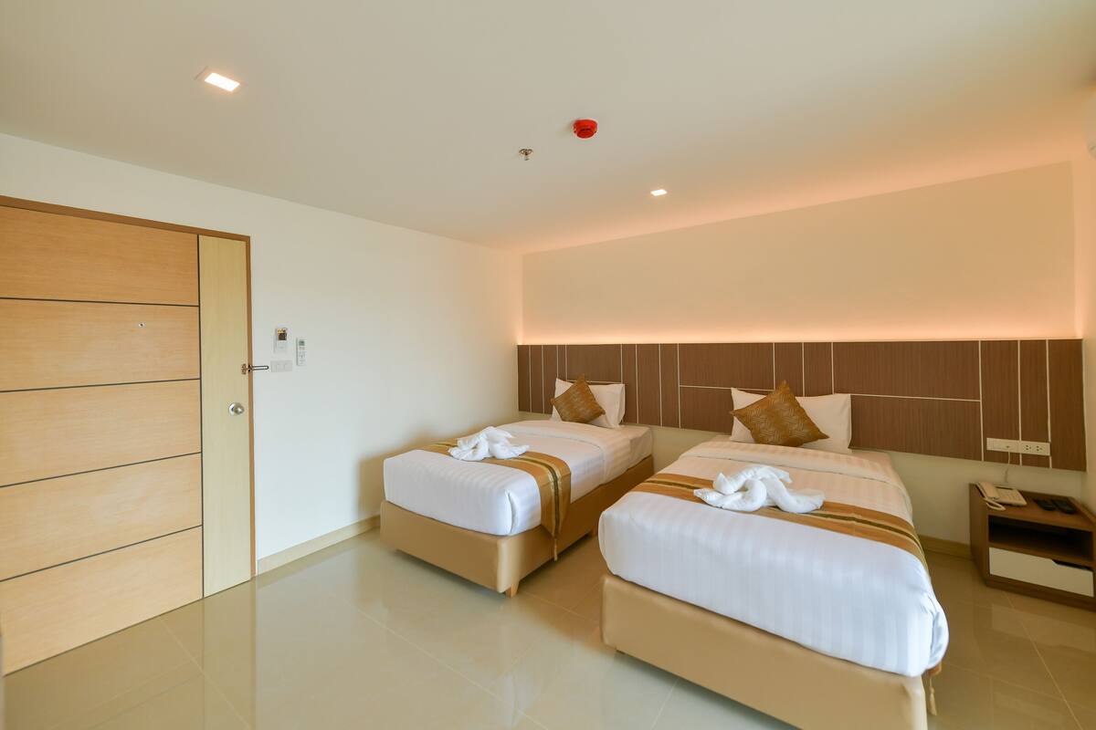 A hotel room with two beds, a wooden door, a nightstand with a phone, and a wall-mounted light fixture.