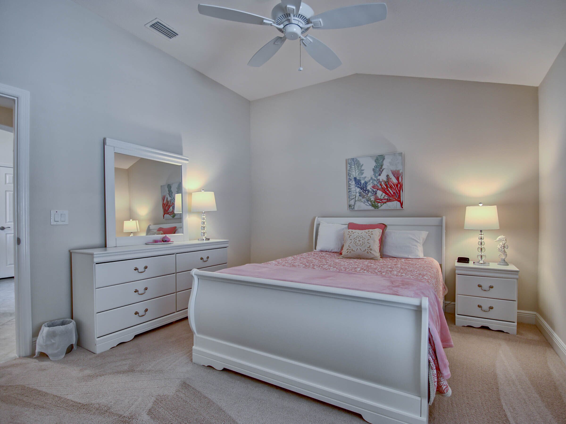 A bedroom with a white bed, a ceiling fan, a nightstand with a lamp, a dresser, and a mirror.