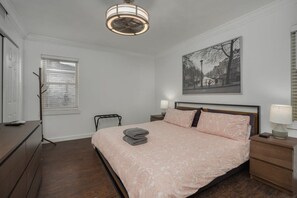 A bedroom with a bed, bedside tables, a nightstand, a window with blinds, a wall-mounted picture, and a ceiling fan.