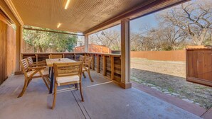 A covered patio area with a wooden table and chairs, large windows, and a view of a backyard.