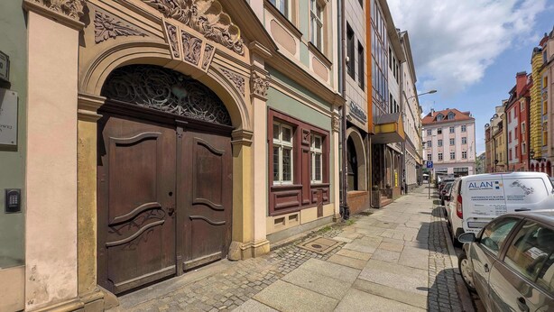Une rue bordée d’immeubles, une porte d’entrée en bois à deux battants et des voitures garées.
