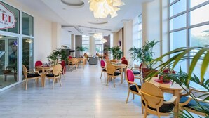 Eine großzügige Hotelhalle mit mehreren Essplätzen, Tischen und Stühlen, großen Fenstern und Zimmerpflanzen.