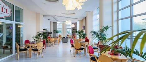 Un amplio vestíbulo de hotel con varias mesas y sillas para comer, ventanas grandes y plantas de interior.