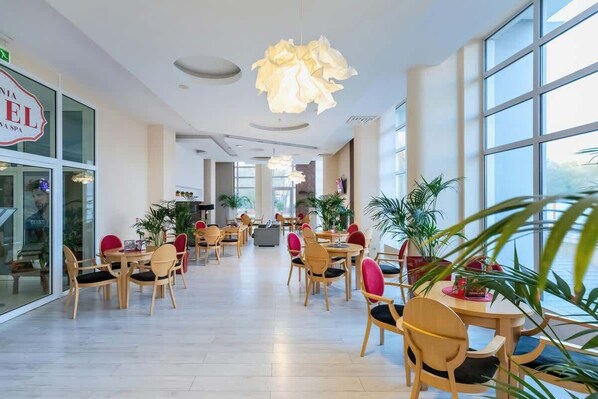 Un amplio vestíbulo de hotel con varias mesas y sillas para comer, ventanas grandes y plantas de interior.