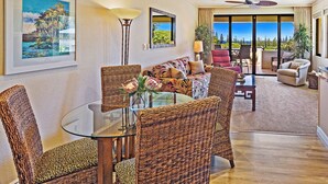 A spacious living area with a glass dining table surrounded by wicker chairs, a sofa with tropical prints, and a view of the outdoors through large windows.